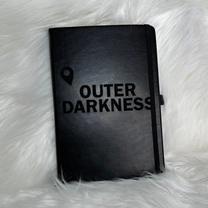 May include: A black leather-bound journal with the words "OUTER DARKNESS" embossed on the front cover. A small pin icon is also embossed on the cover.
