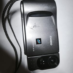 May include: A dark grey Casio QV-10 digital camera with a black wrist strap. The front of the device features the Casio logo, a small colourful icon, and the text LCD DIGITAL CAMERA QV-10. The camera has a rotating lens section on the bottom right.