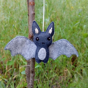 May include: A gray felt bat with white felt ears and a white felt belly. The bat is hanging from a white string.