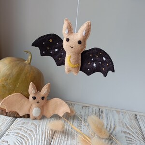 May include: Two felt bat ornaments, one hanging and one lying on a wooden surface. The hanging bat has black wings with gold stars and a crescent moon, and the lying bat is a light brown color.