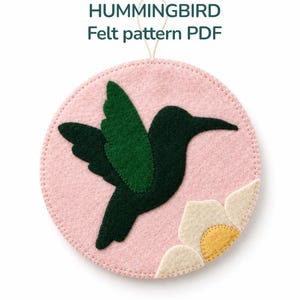 May include: A pink felt hummingbird pattern with a dark green hummingbird silhouette and a white and yellow flower. The text "HUMMINGBIRD Felt pattern PDF" is at the top. A loop is attached for hanging.