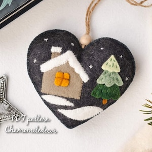Peut inclure: Ornement de Noël en forme de cœur en feutre gris foncé, orné d'une maison et d'un arbre. L'ornement présente des détails blancs, une maison beige et un arbre vert. Il est suspendu à une ficelle. Le texte sur l'ornement indique "PDF pattern Chamomiledecor".