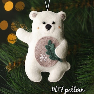 May include: A white felt polar bear ornament with a green leaf and a snowflake on its belly. The ornament is hanging on a Christmas tree. The text "PDF pattern Chamomiledecor" is at the bottom of the image.