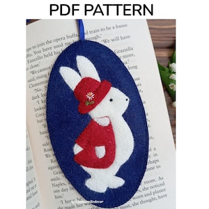 Puede incluir: Marcapáginas de fieltro hecho a mano en forma ovalada, con un conejito blanco con un atuendo y sombrero rojos, sobre un fondo azul oscuro. El texto "Felt bookmark PDF PATTERN" está en la parte superior. El marcapáginas está en un libro abierto.