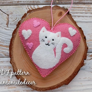 A pink felt heart with a white felt cat inside. The cat has a curled tail and is looking to the right. The heart has white felt hearts around the cat. The heart is hanging from a string and is on a wooden background. The text 'PDF pattern Chamomiledecor' is at the bottom of the image.