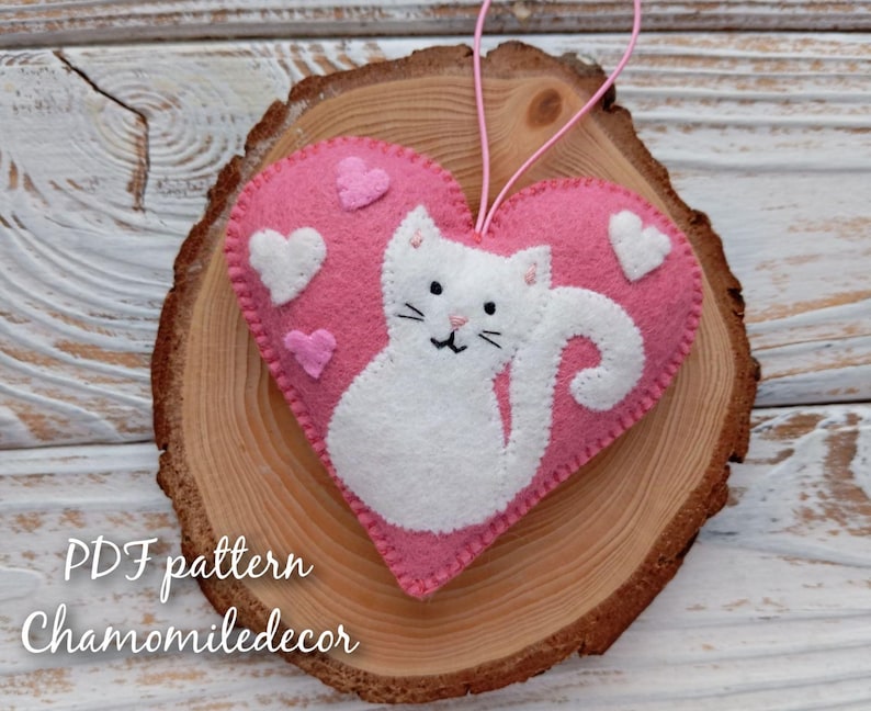 A pink felt heart with a white felt cat inside. The cat has a curled tail and is looking to the right. The heart has white felt hearts around the cat. The heart is hanging from a string and is on a wooden background. The text 'PDF pattern Chamomiledecor' is at the bottom of the image.