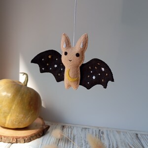 May include: A brown felt bat with black wings and a yellow crescent moon embroidered on its chest. The wings are decorated with white and gold stars.