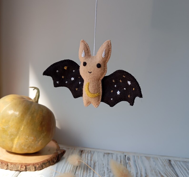 May include: A brown felt bat with black wings and a yellow crescent moon embroidered on its chest. The wings are decorated with white and gold stars.