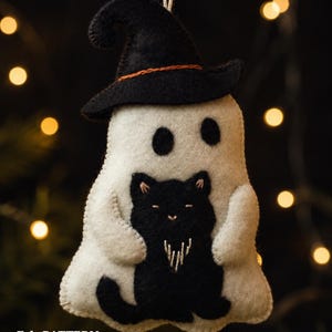 Könnte beinhalten: Ein weißes Filz-Geisterornament mit einem schwarzen Hexenhut mit orangefarbenem Band, das eine kleine schwarze Katze hält. Der Geist hat schwarze gestickte Augen und die Katze zeigt weiße Stickdetails. Unten steht der Text: Felt PATTERN By Chamomiledecor.