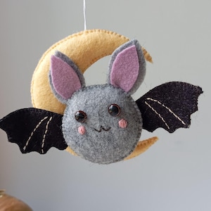 May include: A gray felt bat with pink ears and black wings hanging from a yellow felt crescent moon. The bat has embroidered eyes and a stitched smile.