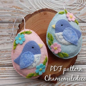 Peut inclure: Deux ornements d'oiseaux en feutre en forme d'œufs. L'un est rose avec un oiseau bleu et l'autre est bleu avec un oiseau bleu. Les deux ont des détails floraux et sont suspendus à une ficelle blanche.