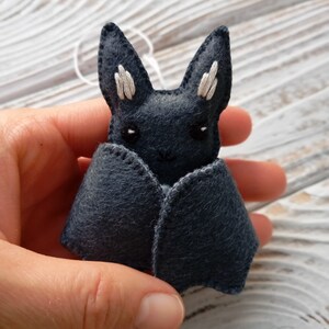 May include: A small, gray felt bat with white embroidered ears and eyes. The bat is held in a hand.