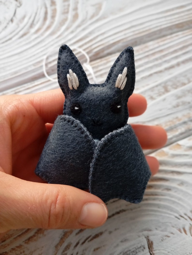 May include: A small, gray felt bat with white embroidered ears and eyes. The bat is held in a hand.