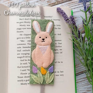 May include: A felt bunny bookmark with a pink body, white ears, and a green background. The bunny is sitting on a bed of green felt with yellow, purple, and pink flowers. The bookmark is attached to a grey ribbon.