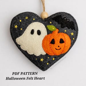 May include: A heart-shaped dark gray felt ornament featuring a white ghost, an orange jack-o'-lantern, and a black bat. The design includes small yellow embroidered stars and a wooden bead at the top for hanging. Text reads PDF PATTERN Halloween Felt Heart.