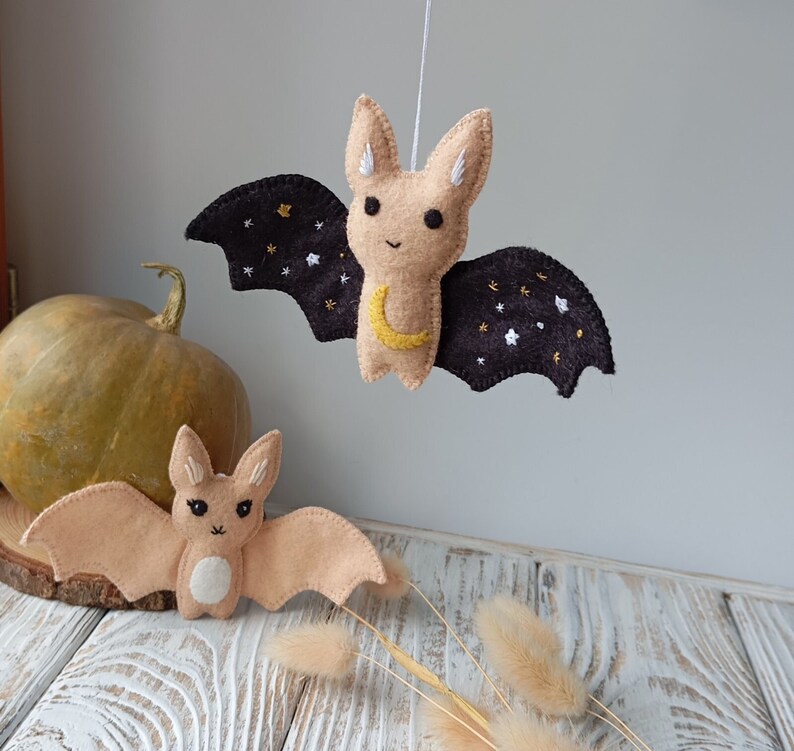 May include: Two felt bat ornaments, one hanging and one lying on a wooden surface. The hanging bat has black wings with gold stars and a crescent moon, and the lying bat is a light brown color.