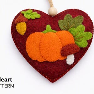 Pode incluir: Um ornamento de feltro em forma de coração vermelho escuro com uma abóbora laranja, uma bolota amarela com chapéu castanho, folhas verdes e um pequeno cogumelo com chapéu castanho e caule branco. Uma conta de madeira clara está presa no topo.