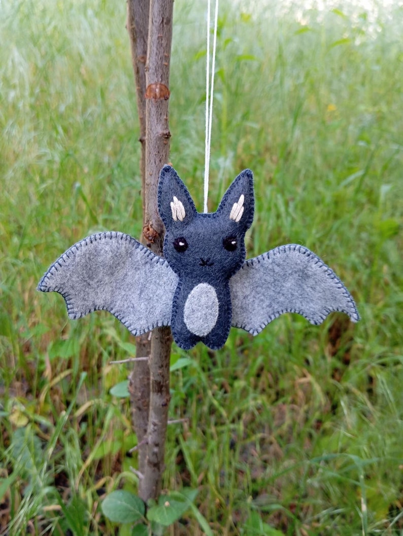 May include: A gray felt bat with white felt ears and a white felt belly. The bat is hanging from a white string.