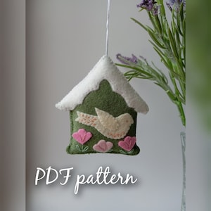 Può includere: Un ornamento per la casa a forma di casetta per uccelli in feltro con un uccello bianco, fiori rosa e un tetto verde con una cima bianca innevata. L'ornamento è appeso a uno spago bianco. Il testo "PDF pattern" è scritto sotto l'ornamento.