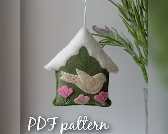 May include: A felt birdhouse ornament with a white bird, pink flowers, and a green roof with a white snowy top. The ornament is hanging from a white string. The text "PDF pattern" is written below the ornament.