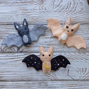 May include: Three felt bat ornaments hanging from white string. The bats are gray, peach, and black with embroidered details. The black bat has a crescent moon and stars on its wings.