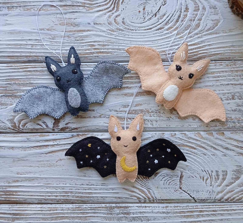 May include: Three felt bat ornaments hanging from white string. The bats are gray, peach, and black with embroidered details. The black bat has a crescent moon and stars on its wings.