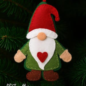 May include: A handmade Christmas gnome ornament with a red hat, white beard, and green coat. The gnome has a red heart on its chest and tan mittens. The ornament is hanging from a red string. Text on the image reads "PDF pattern Chamomiledecor".