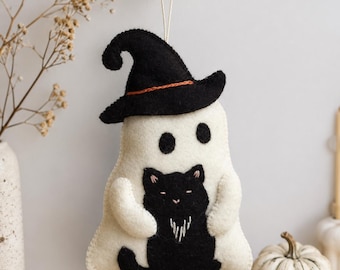 May include: A felt hanging ornament shaped like a white ghost wearing a black witch hat with an orange band, holding a small black cat. The ghost has two black oval eyes and the cat has embroidered details. Text reads PDF PATTERN By Chamomiledecor.