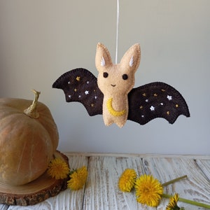 May include: A brown felt bat with a yellow crescent moon and white stars embroidered on its wings. The bat is hanging from a string and has a beige face with black eyes.