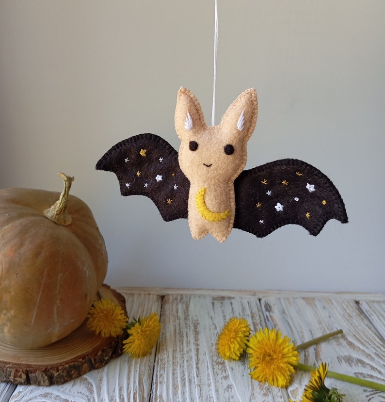 May include: A brown felt bat with a yellow crescent moon and white stars embroidered on its wings. The bat is hanging from a string and has a beige face with black eyes.