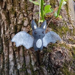 May include: A gray felt bat with white eyes and a white belly hanging from a string. The bat is hanging against a tree trunk.