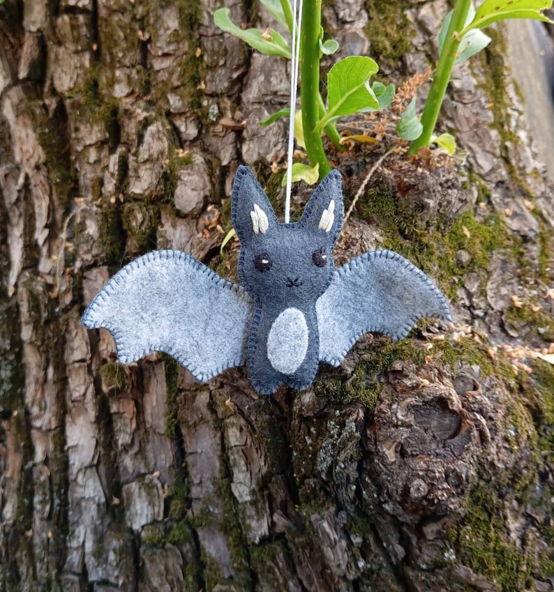 May include: A gray felt bat with white eyes and a white belly hanging from a string. The bat is hanging against a tree trunk.