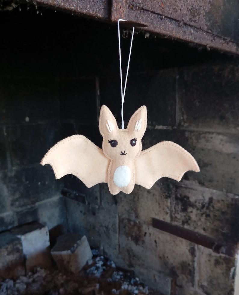 May include: A beige felt bat ornament with embroidered eyes and a white felt belly. The bat is hanging from a white string.