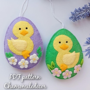 Könnte beinhalten: Zwei Filz-Ostereier-Anhänger mit einem gelben Küken-Design. Die Eier sind mit weißen Blumen und grünen Blättern verziert. Die Eier hängen an weißen Schnüren. Der Text "PDF pattern Chamomiledecor" befindet sich am unteren Rand des Bildes.