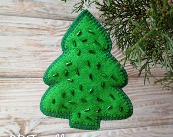Könnte beinhalten: Ein grüner Filz-Weihnachtsbaum-Anhänger mit grünen Perlen, die darauf genäht sind. Der Anhänger hängt an einer weißen Schnur vor einem weißen Holz-Hintergrund. Der Text "PDF pattern" steht unter dem Anhänger.