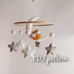 Puede incluir: Un móvil de bebé de fieltro con una luna creciente, un zorro durmiendo y estrellas. El móvil está colgado de un anillo de madera. El texto "PDF pattern" es visible en la imagen.
