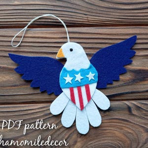 May include: A felt craft pattern for a patriotic eagle with blue wings, a white body, and a red, white, and blue star-spangled banner on its chest. The pattern is available as a PDF download from Chamomiledecor.