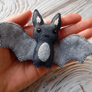 May include: A gray felt bat with white stitching and black eyes. The bat has a white felt belly and is a handmade ornament.