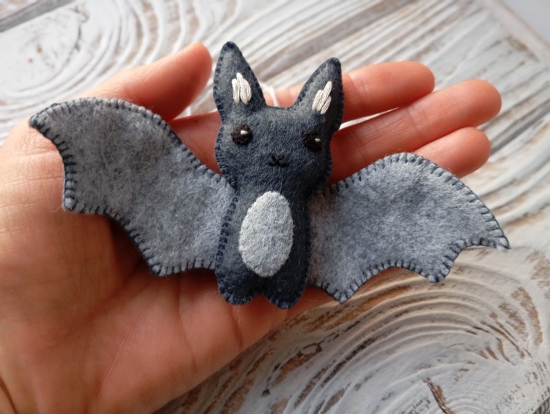 May include: A gray felt bat with white stitching and black eyes. The bat has a white felt belly and is a handmade ornament.