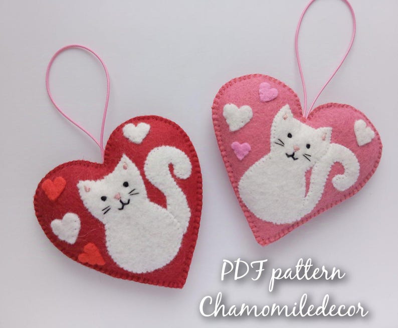 Two felt heart ornaments with white cat designs. The heart on the left is red with white hearts and the heart on the right is pink with white hearts. The text 'PDF pattern Chamomiledecor' is at the bottom of the image.