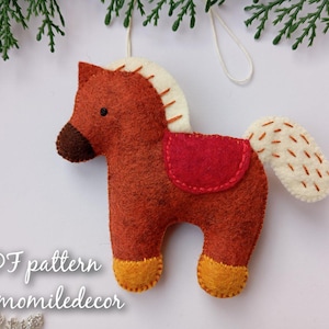 Puede incluir: Adorno de caballo de fieltro hecho a mano en tonos marrón, rojo y crema. El caballo tiene una silla de montar roja, crin y cola color crema y cascos amarillos. El texto "PDF pattern Chamomiledecor" es visible.