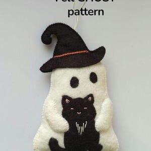 Op de afbeelding: Een witte vilten geest met een zwarte heksenhoed en een zwart kattenontwerp. De geest heeft twee zwarte ogen en houdt de zwarte kat vast. De tekst "Felt GHOST pattern" staat bovenaan.