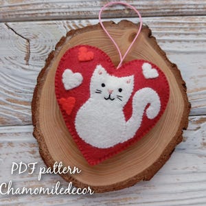 A red felt heart with a white felt cat inside. The heart has white felt hearts around the cat. The cat has a pink nose and whiskers. The heart is on a wooden surface. PDF pattern Chamomiledecor