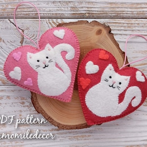 Two felt heart ornaments with white cat designs and pink and red hearts. The ornaments have pink string loops for hanging. The text 'PDF pattern chamomiledecor' is at the bottom of the image.