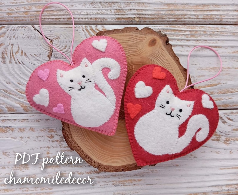 Two felt heart ornaments with white cat designs and pink and red hearts. The ornaments have pink string loops for hanging. The text 'PDF pattern chamomiledecor' is at the bottom of the image.
