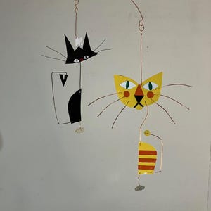 以下が含まれることがあります： 2匹の様式化された猫のフィギュアを特徴とする気まぐれな猫のモビール。1つは白いアクセントのある黒、もう1つはオレンジとグリーンのディテールのある黄色です。銅線とチェーンで吊り下げられています。