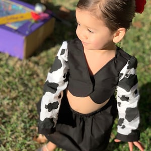 May include: A young girl wearing a black and white cow print top and skirt. The top is a cropped style with a black collar and the skirt is a simple black skirt. The girl is wearing silver shoes and has a red flower in her hair.