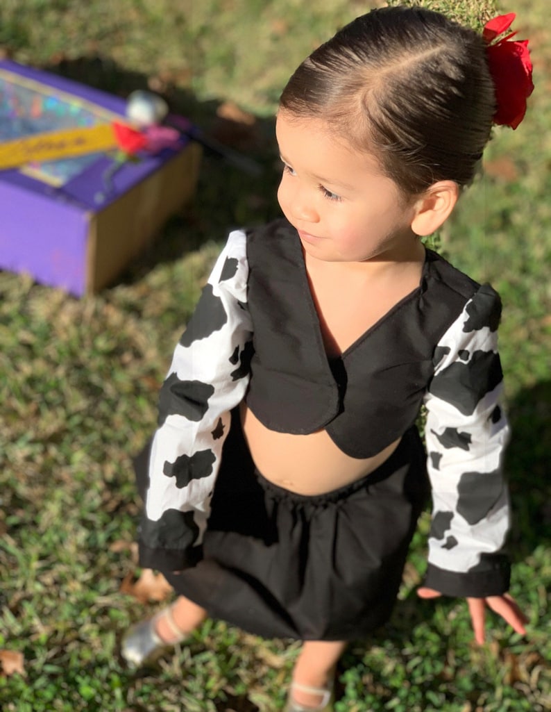 May include: A young girl wearing a black and white cow print top and skirt. The top is a cropped style with a black collar and the skirt is a simple black skirt. The girl is wearing silver shoes and has a red flower in her hair.