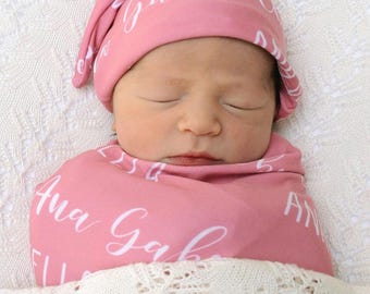 May include: A pink baby swaddle blanket with the name "Ana Gabriela" written in white cursive. A pink baby hat with the name "Gabriela" written in white cursive is on top of the baby's head.