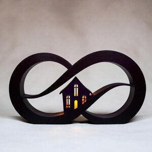 Op de afbeelding: Een donkerbruin houten oneindigheidssymbool met een klein huis silhouet in het midden. Het huis heeft verlichte ramen, waardoor een warme gloed ontstaat. Het ontwerp is een decoratief stuk, mogelijk een kaarsenhouder of een nachtlampje.